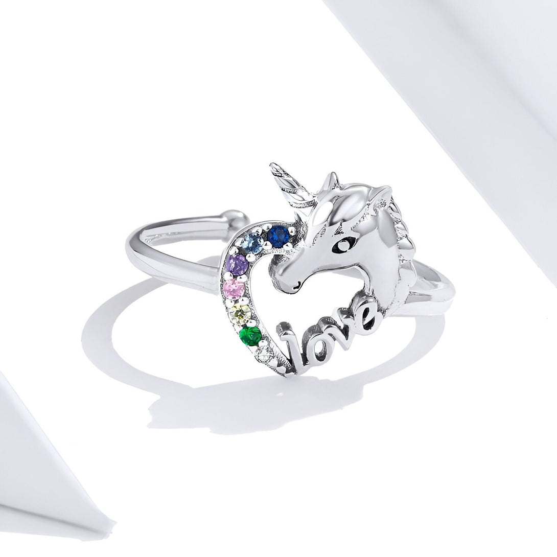 Une bague en argent avec un cœur et une licorne. La licorne est sertie de pierres précieuses multicolores et le mot "love" est écrit en lettres minuscules sur le cœur.