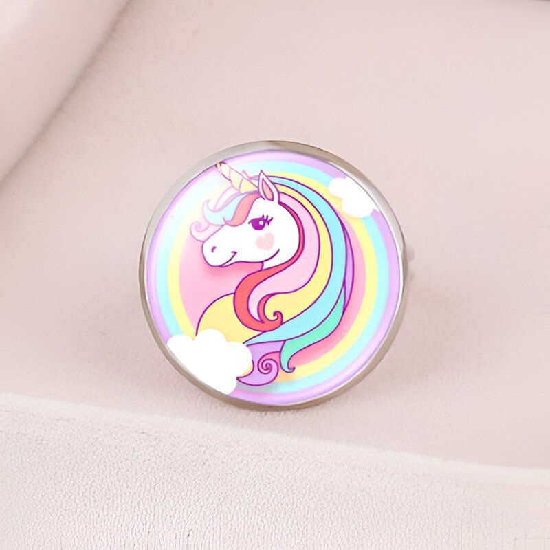 L'image représente une bague en métal avec un cabochon en verre rond. Le cabochon est décoré d'une image d'une licorne blanche avec une crinière et une queue arc-en-ciel. La licorne est entourée de nuages blancs et d'un arc-en-ciel.