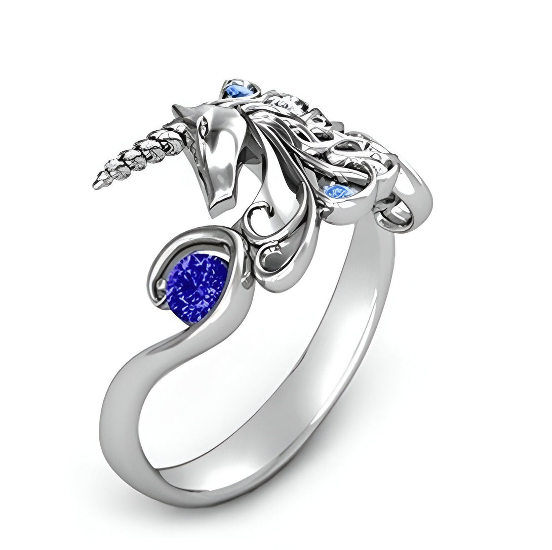 Une bague en argent avec une pierre précieuse bleue et des accents de pierres précieuses bleues plus petites. La bague est en forme de licorne avec la tête de la licorne tournée vers le haut et la queue enroulée autour de son corps.