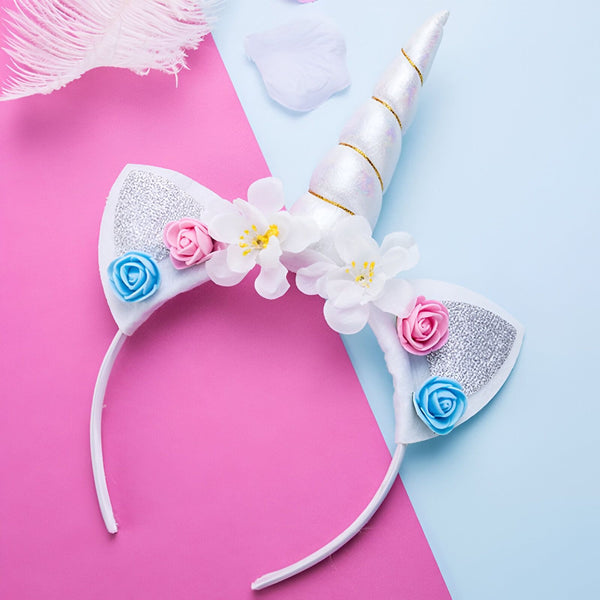 Serre-tête licorne blanc avec corne argentée, oreilles scintillantes, et fleurs roses, bleues et blanches, sur fond rose et bleu. Accessoire de déguisement féérique., 45501339664639 | Licorne Shop