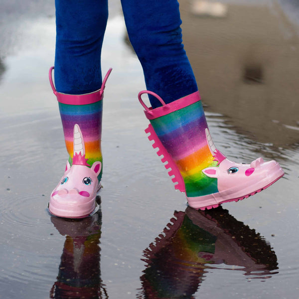 Enfant portant des bottes de pluie arc-en-ciel à motif de licorne dans une flaque d'eau. Bottes roses avec des détails de licorne et des poignées roses. Pantalon bleu. Reflet dans l'eau., 45501218652415,45501218685183,45501218717951,45501218750719,45501218783487,45501218816255,45501218849023,45501218881791,45501218914559 | Licorne Shop