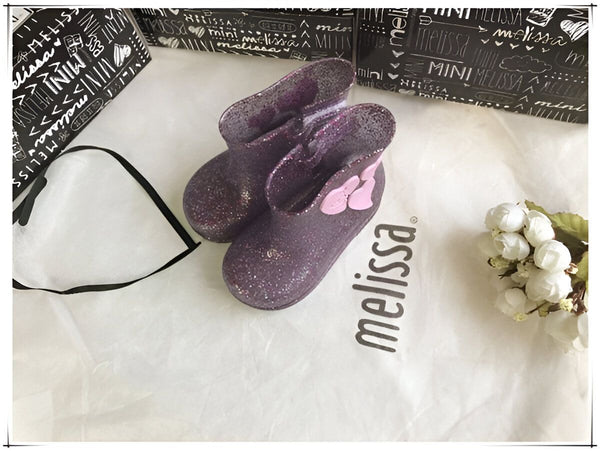 Bottes de pluie violettes à paillettes Mini Melissa sur sac en tissu blanc avec logo Melissa, emballage et fleurs blanches. Chaussures pour enfant tendance et mignonnes., 45501102358783,45501102391551,45501102424319,45501102457087,45501102489855,45501102522623,45501102555391,45501102588159,45501102620927 | Licorne Shop