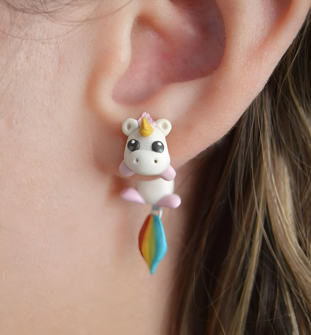 Une image d'une boucle d'oreille en forme de licorne blanche avec une crinière et une queue arc-en-ciel. La licorne est suspendue à l'oreille par un fil d'argent.