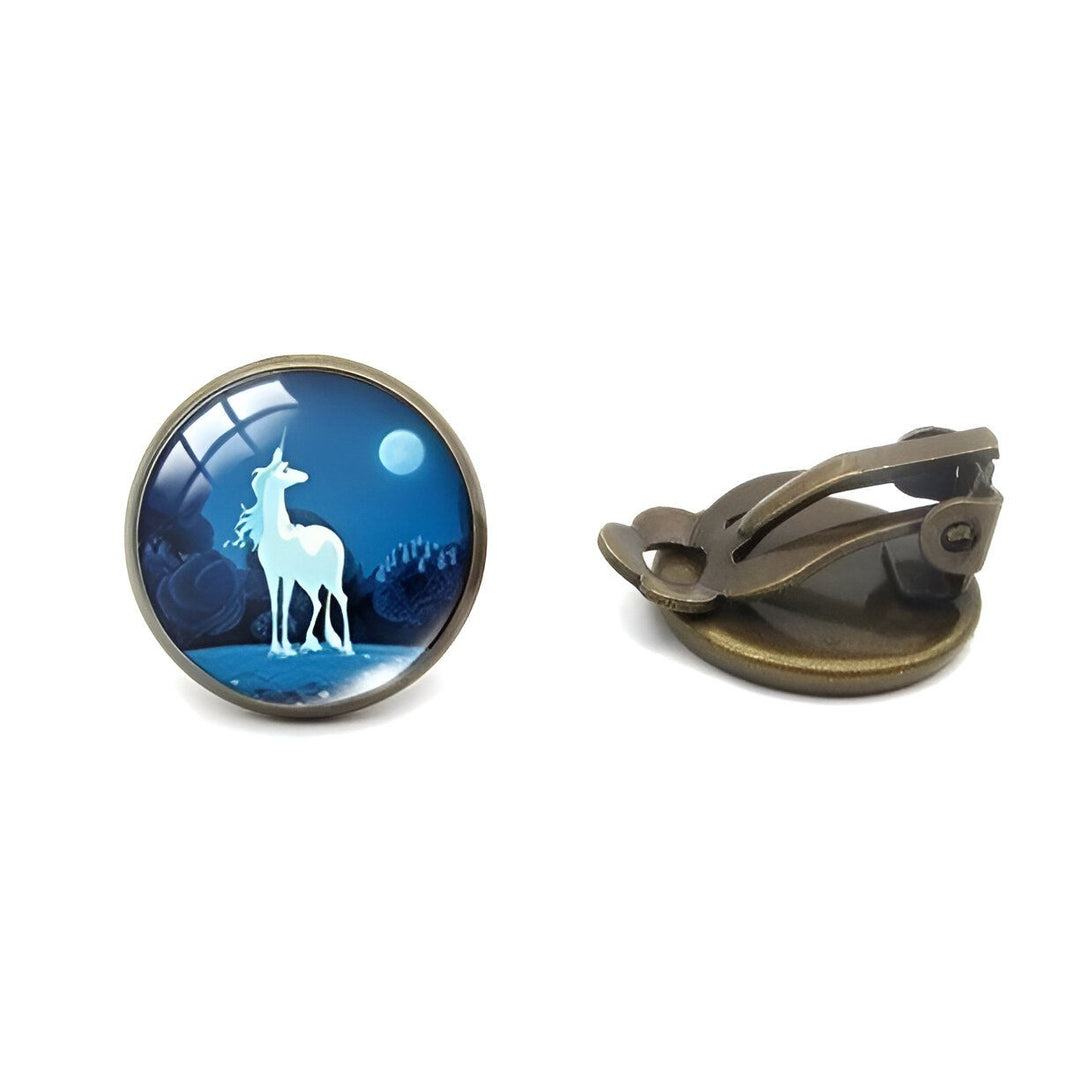 Une paire de boucles d'oreilles en bronze avec un cabochon en verre rond. Le cabochon représente une illustration d'une licorne blanche se tenant devant une pleine lune. La licorne est entourée d'arbres et de montagnes. Le fond est bleu nuit.