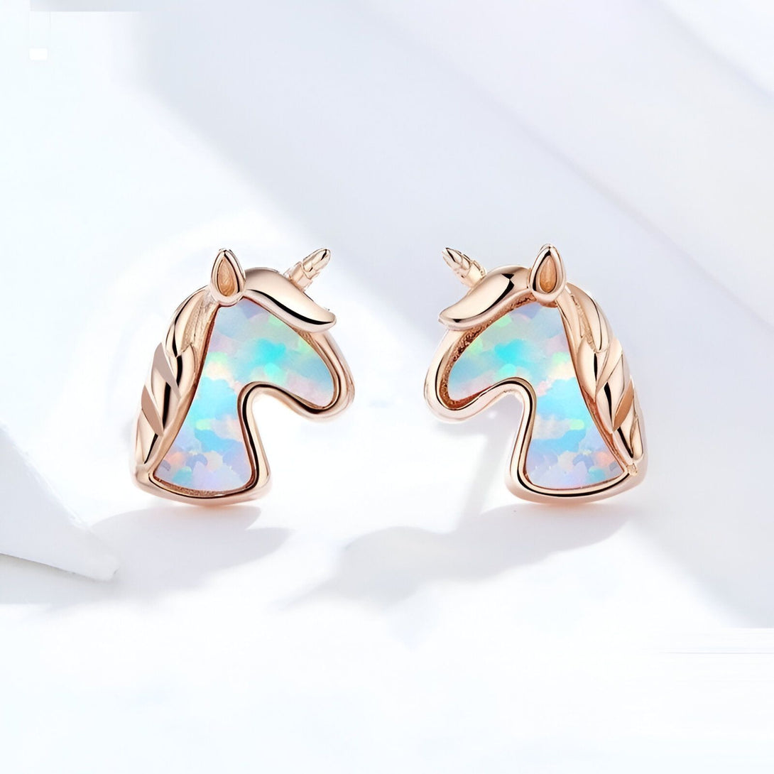 Une paire de boucles d'oreilles en or rose en forme de licorne. Les licornes ont une crinière et une queue en opale blanche.