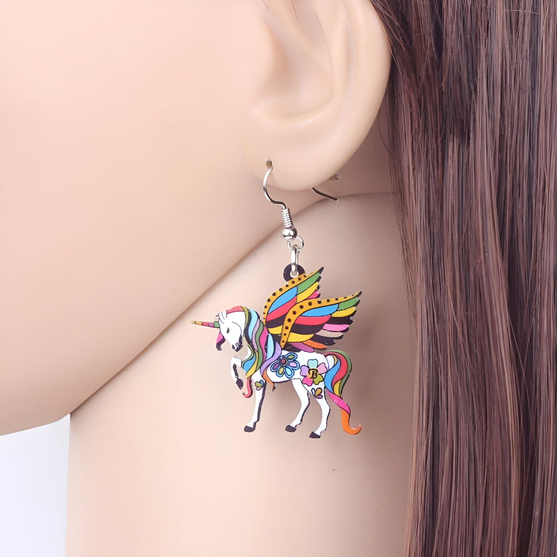 Une image d'une boucle d'oreille en forme de licorne. La licorne est blanche avec une crinière et une queue arc-en-ciel. Elle a des ailes multicolores et une corne dorée. La boucle d'oreille est en métal argenté.