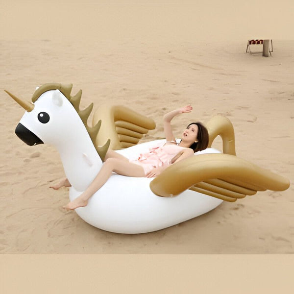 Une femme se détend sur une bouée licorne gonflable sur une plage de sable clair. La licorne est blanche avec une crinière et des ailes dorées. La femme, en combinaison rose, lève un bras., 45501242867967 | Licorne Shop