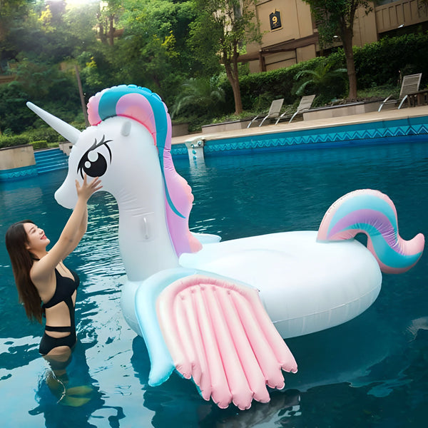 Une femme en maillot de bain noir dans une piscine touche une grande bouée gonflable licorne blanche avec des accents roses et bleus. La piscine est entourée de végétation., 45501251354879 | Licorne Shop