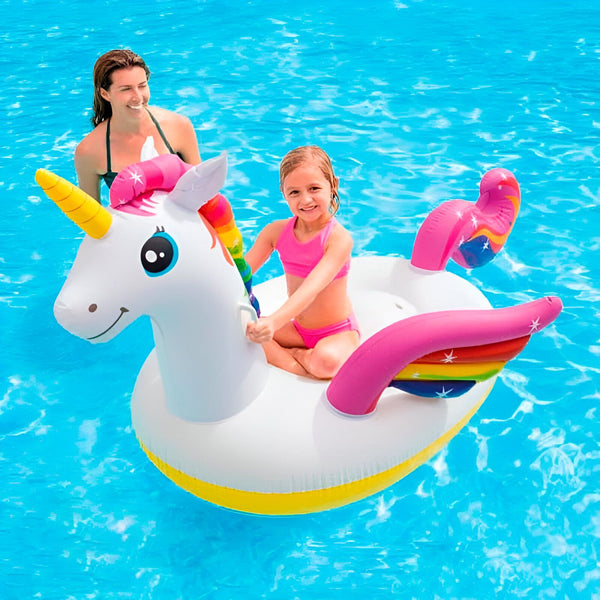 Une fillette assise sur une bouée gonflable en forme de licorne dans une piscine, avec une femme en arrière-plan. La licorne est blanche avec une crinière arc-en-ciel et des ailes roses., 45501250273535 | Licorne Shop