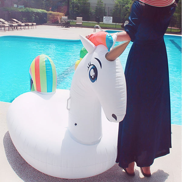 Femme près d'une piscine avec une bouée gonflable licorne blanche à crinière arc-en-ciel. Elle porte une robe bleue foncée et un chapeau rayé., 45501250732287 | Licorne Shop