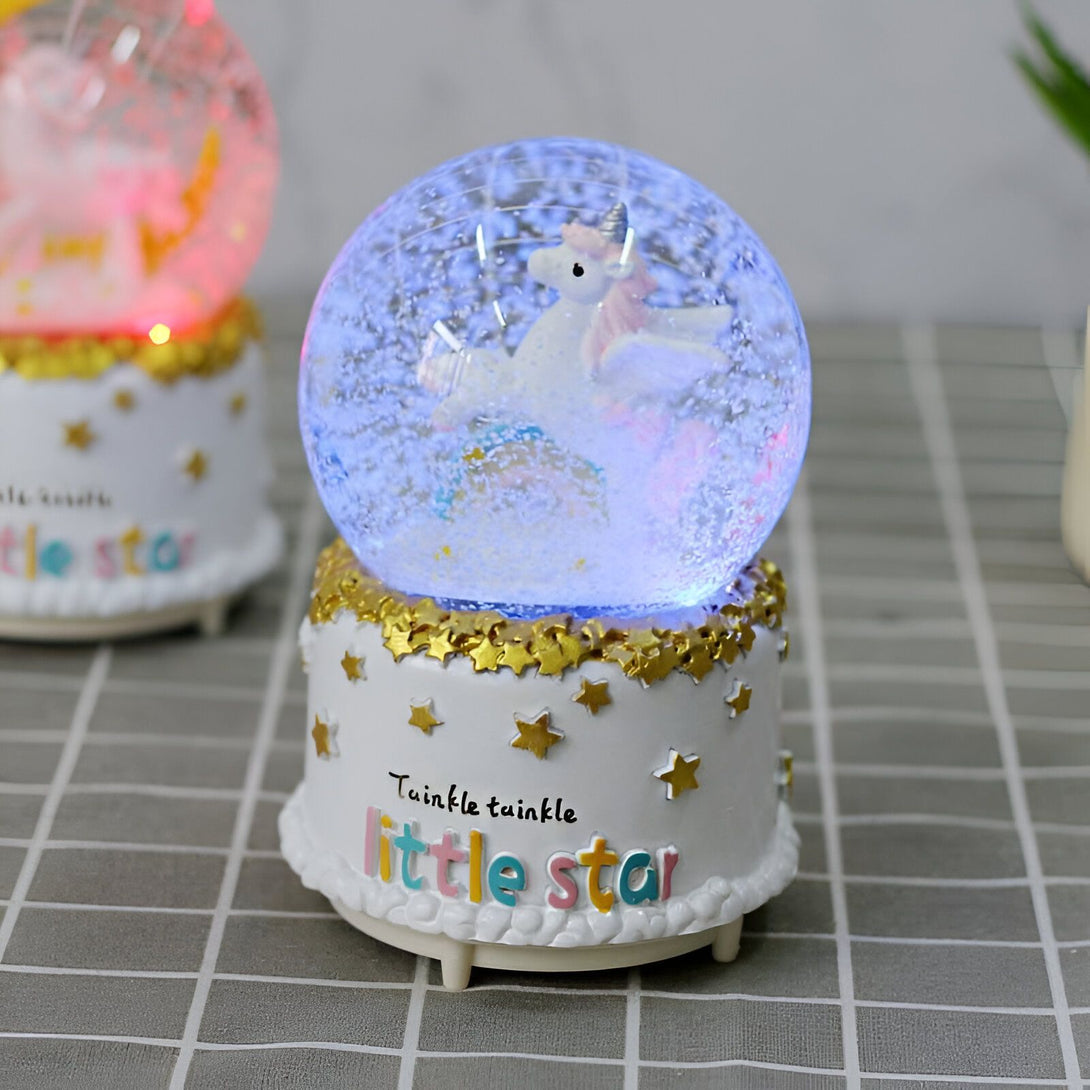 Boule à neige musicale licorne scintillante. Objet de décoration avec une licorne blanche à l'intérieur d'une boule transparente remplie de paillettes, posée sur un socle blanc décoré d'étoiles et de l'inscription "Twinkle twinkle little star"., 45501133816063 | Licorne Shop