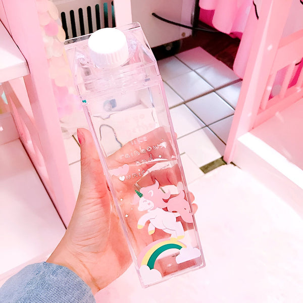 Une main tient une bouteille d'eau transparente en forme de brique de lait avec un bouchon blanc. Des dessins d'arcs-en-ciel et de licornes décorent la bouteille. L'arrière-plan est rose., 45501173858559 | Licorne Shop