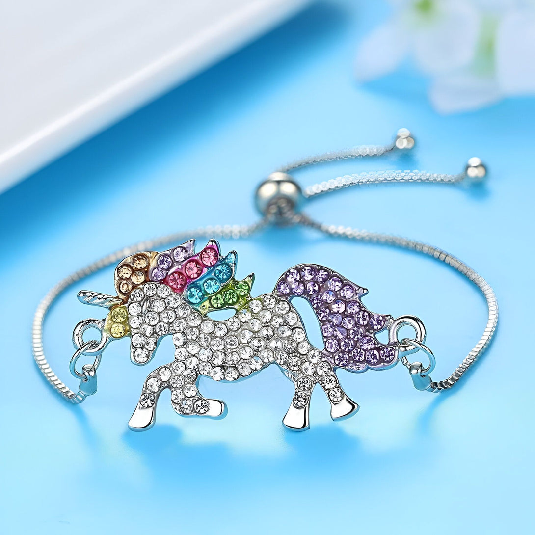 L'image montre un bracelet en argent avec un pendentif en forme de licorne. La licorne est sertie de cristaux de différentes couleurs, notamment du rose, du bleu, du vert et du jaune. Le bracelet est réglable et se ferme à l'aide d'un fermoir mousqueton.