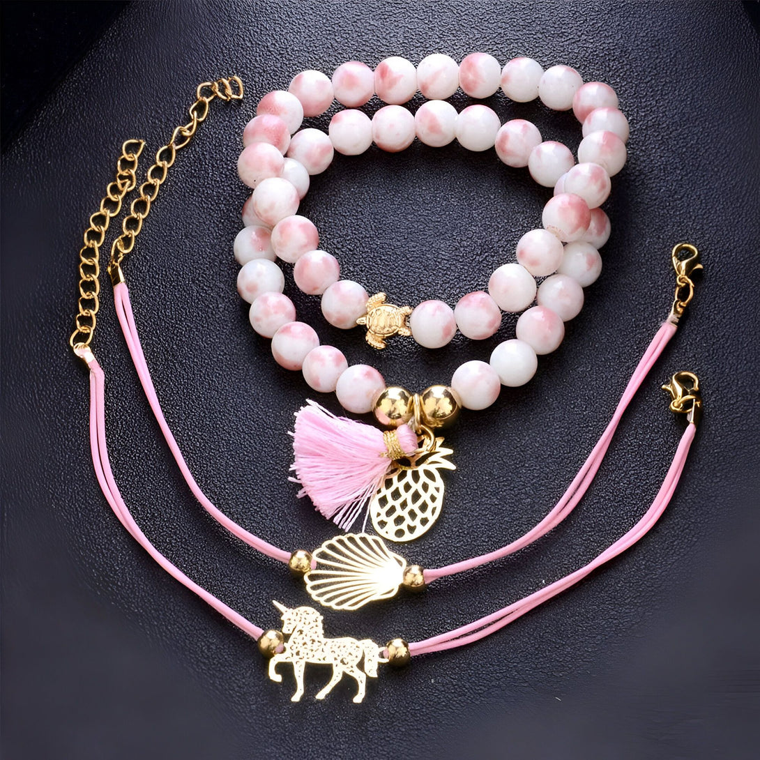 L'image représente un ensemble de trois bracelets. Le premier est un bracelet en perles blanches et roses avec une breloque en forme de tortue en métal doré. Le deuxième est un bracelet en perles blanches et roses avec une breloque en forme de coquillage en métal doré et d'ananas rose . Le troisième est un bracelet en fil rose avec une breloque en forme de licorne en métal doré.