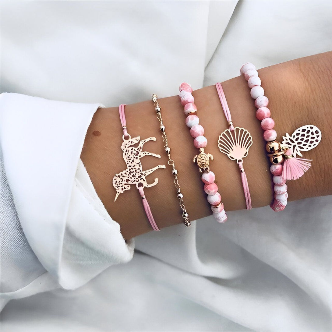 L'image représente cinq bracelets. Il y a un bracelet avec des perles roses et blanches, un bracelet avec des perles en forme de coquillages, un bracelet avec un pendentif en forme de tortue, un bracelet avec un pendentif en forme d'ananas et un bracelet avec un pendentif en forme de licorne.