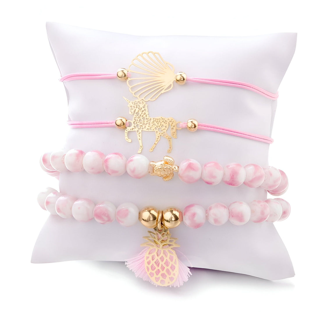 L'image montre trois bracelets en pierre naturelle blanche et rose avec des breloques en forme de coquillage, de licorne, de tortue et d'ananas. Les bracelets sont présentés sur un coussin blanc.