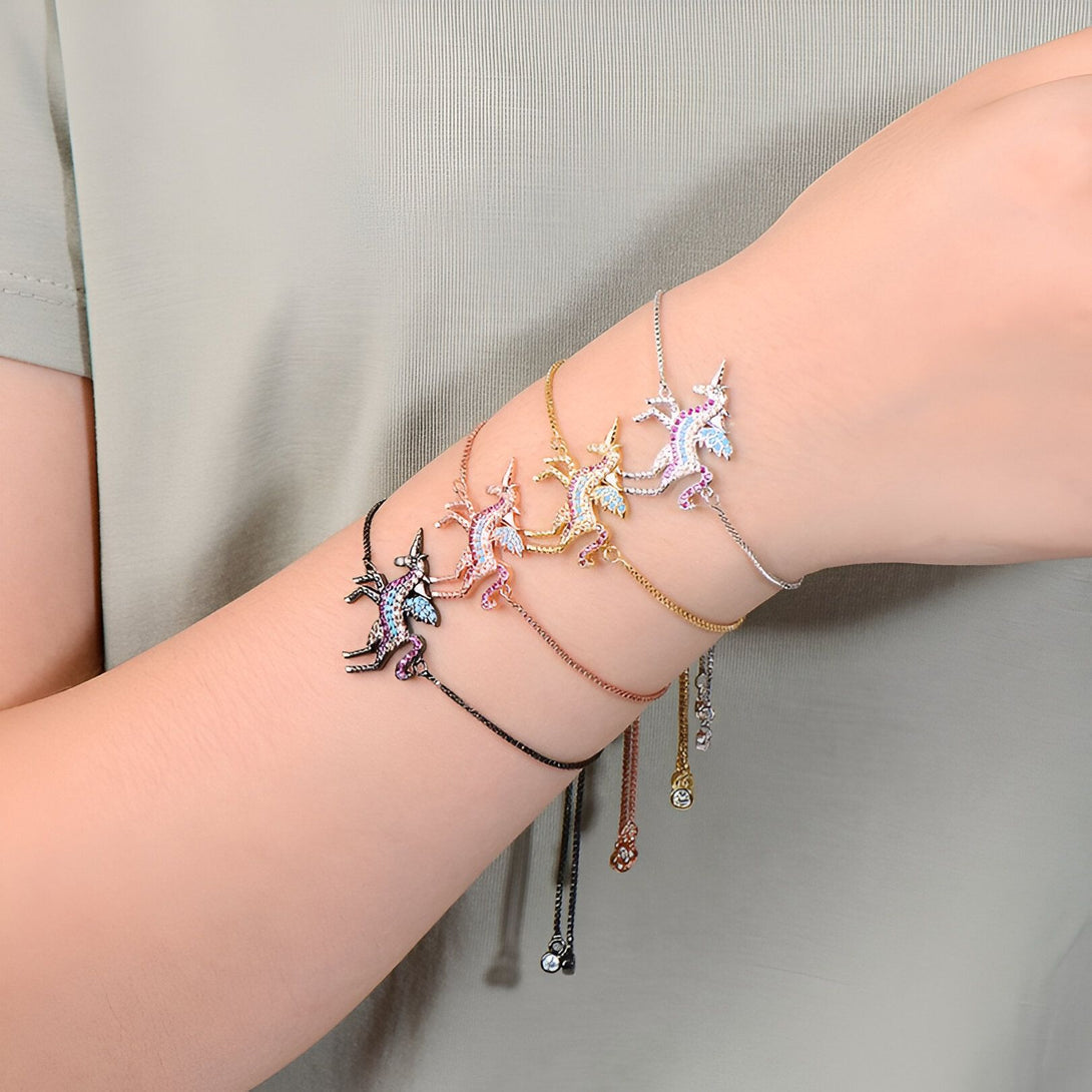 Quatre bracelets avec des pendentifs en forme de licorne sont portés sur le poignet d'une personne. Les licornes sont serties de pierres précieuses colorées. Les bracelets sont en argent, en or rose, en or jaune et en noir.