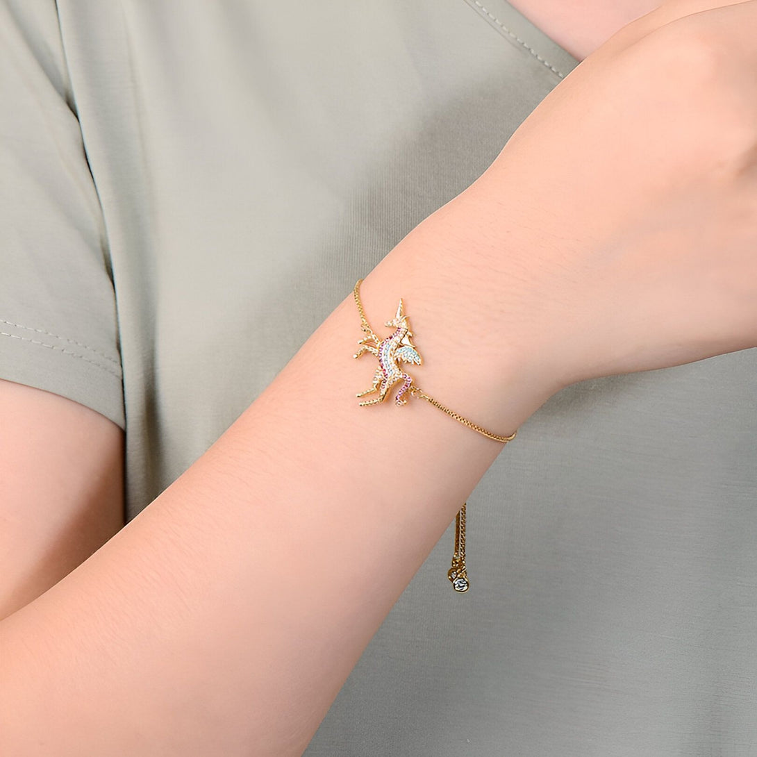 L'image montre un bracelet en or avec un pendentif licorne. La licorne est sertie de pierres précieuses multicolores. Le bracelet est porté par une femme.