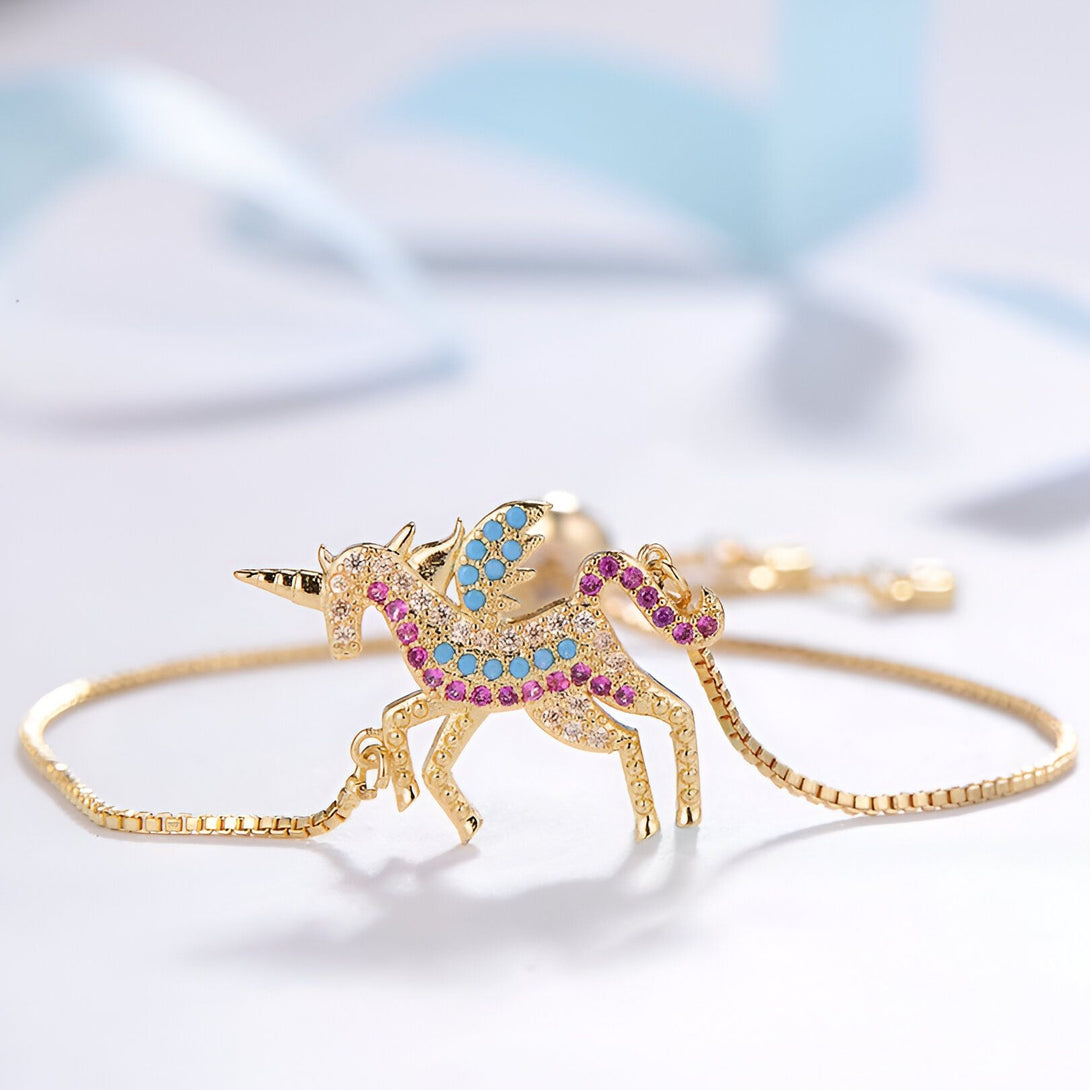 L'image montre un bracelet en or avec un pendentif licorne. La licorne est sertie de pierres précieuses roses, bleues et vertes. Le bracelet est porté sur un poignet de femme.