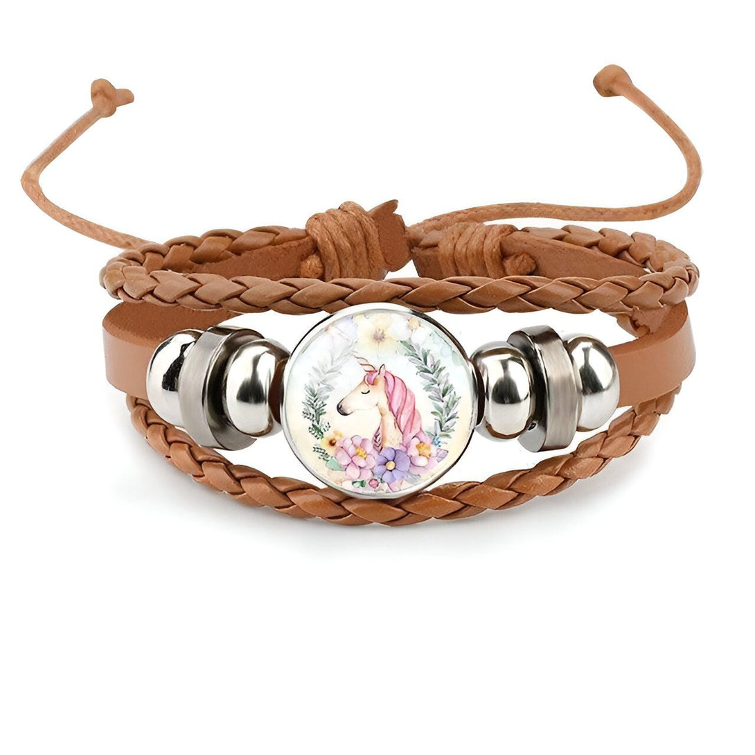 Un bracelet en cuir marron avec un pendentif en verre rond. Le pendentif représente une licorne blanche avec une crinière et une queue roses. La licorne est entourée de fleurs roses et vertes.