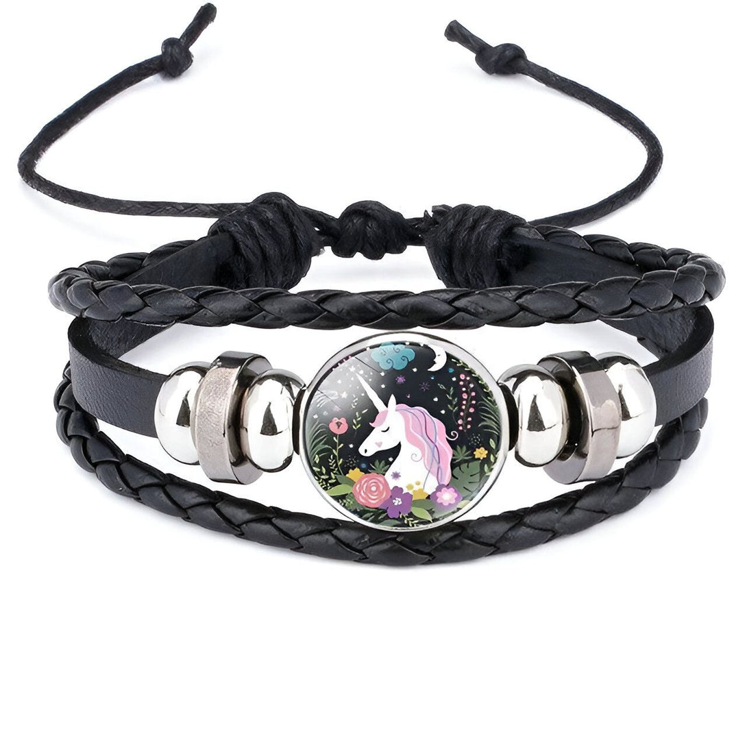 Le bracelet en cuir noir tressé est orné d'une breloque en verre ronde. Le motif de la breloque représente une licorne blanche avec une crinière et une queue roses et violettes. La licorne se tient dans un pré fleuri avec des fleurs roses, violettes et bleues. Le fond est constitué d'un ciel étoilé avec une lune et des nuages.