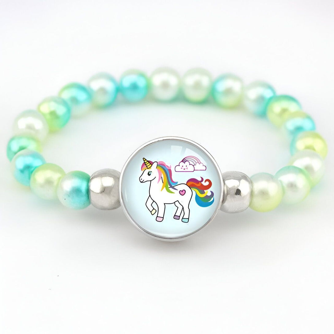 Un bracelet avec une perle centrale en verre qui représente une licorne blanche avec une crinière et une queue arc-en-ciel. La licorne se tient sur un nuage. Le bracelet est fait de perles de couleur vert clair et argenté.