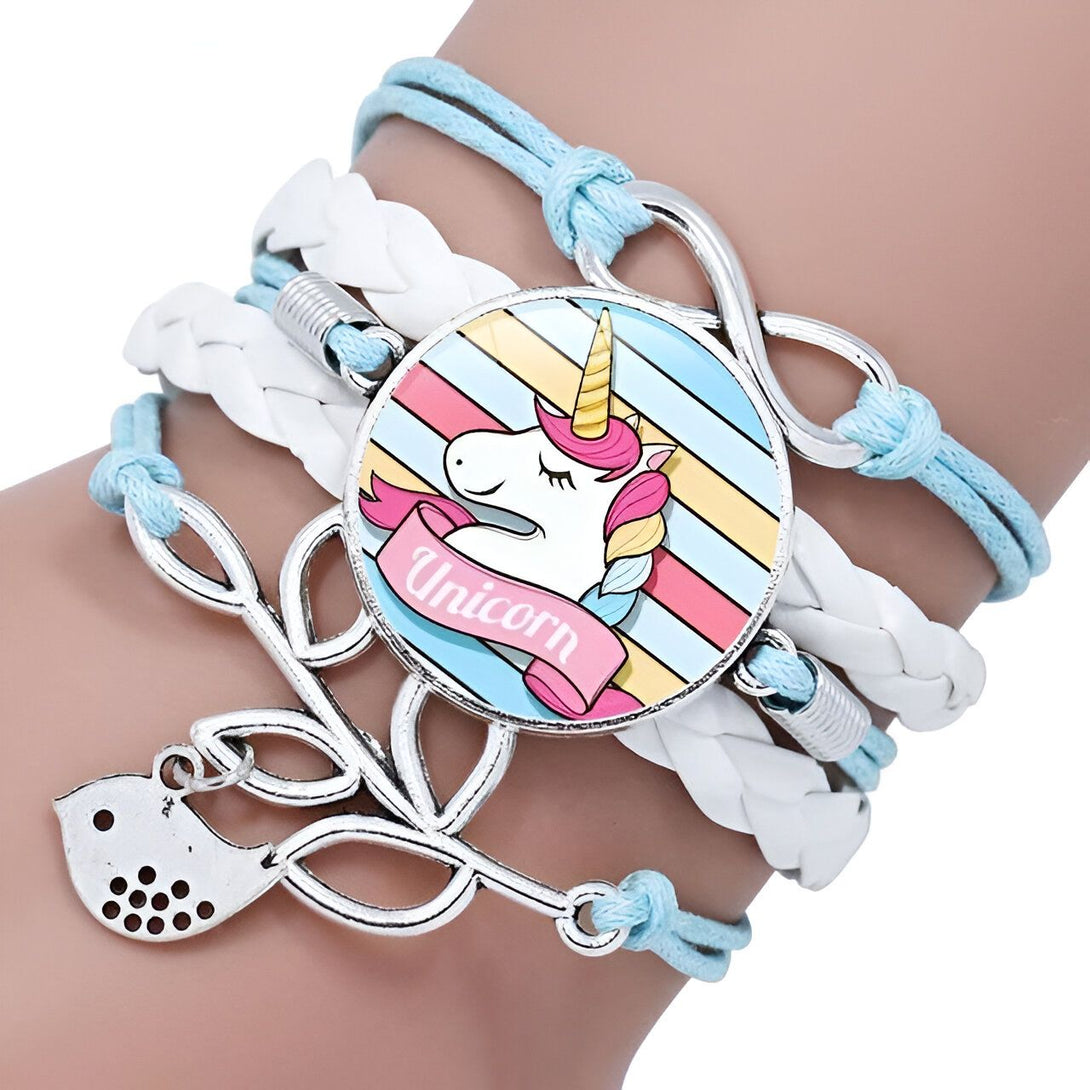 L'image montre un bracelet avec un pendentif en forme de licorne. La licorne est blanche avec une crinière et une queue arc-en-ciel. Le bracelet est en cuir tressé de couleur bleue et blanche. Il est agrémenté de perles en argent et d'un fermoir en forme de nœud.