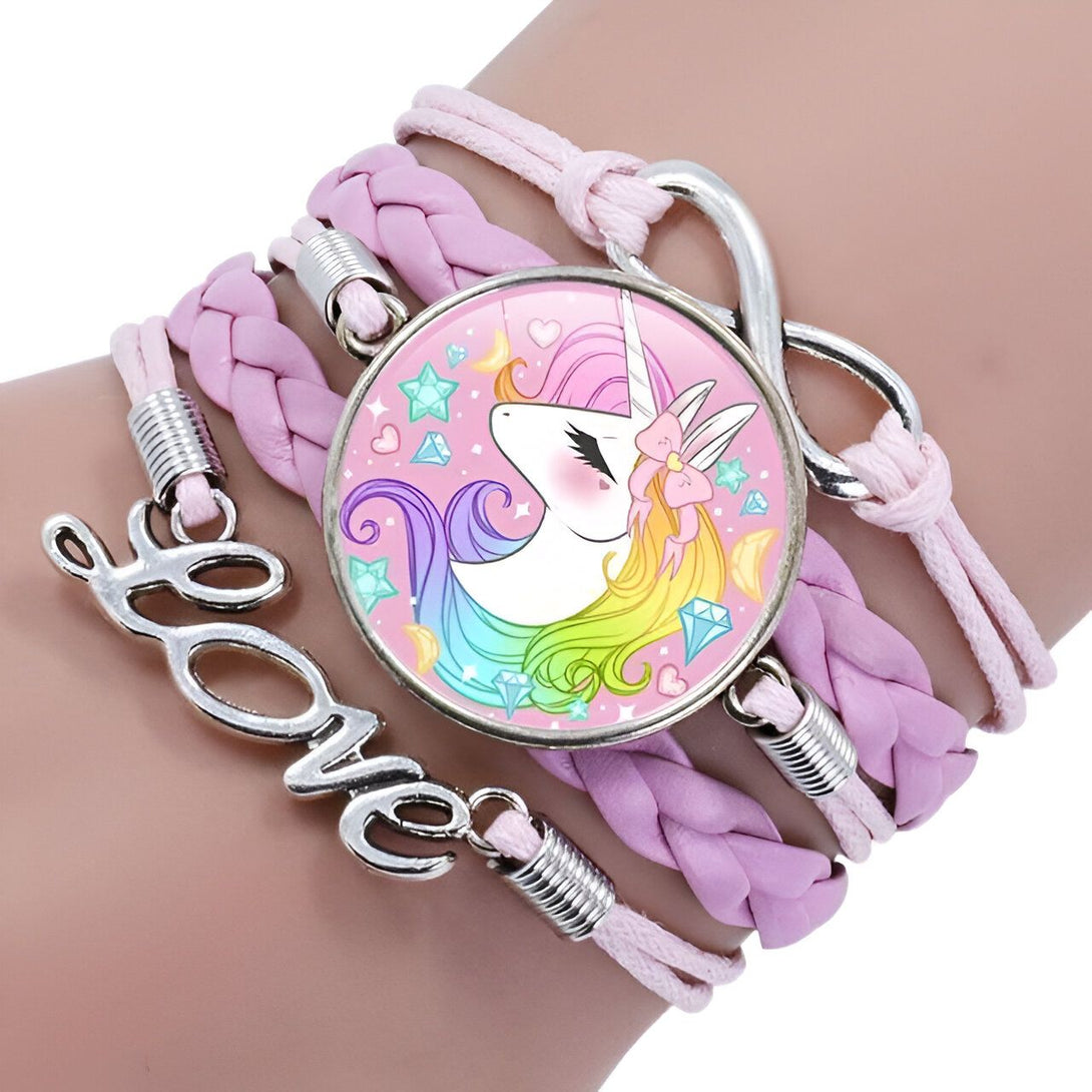 L'image montre un bracelet en cuir tressé rose avec un pendentif en forme de licorne. La licorne est blanche avec une crinière et une queue arc-en-ciel. Elle porte un collier avec un pendentif en forme de cœur. Le bracelet est également décoré de perles en forme d'étoile et de lune.