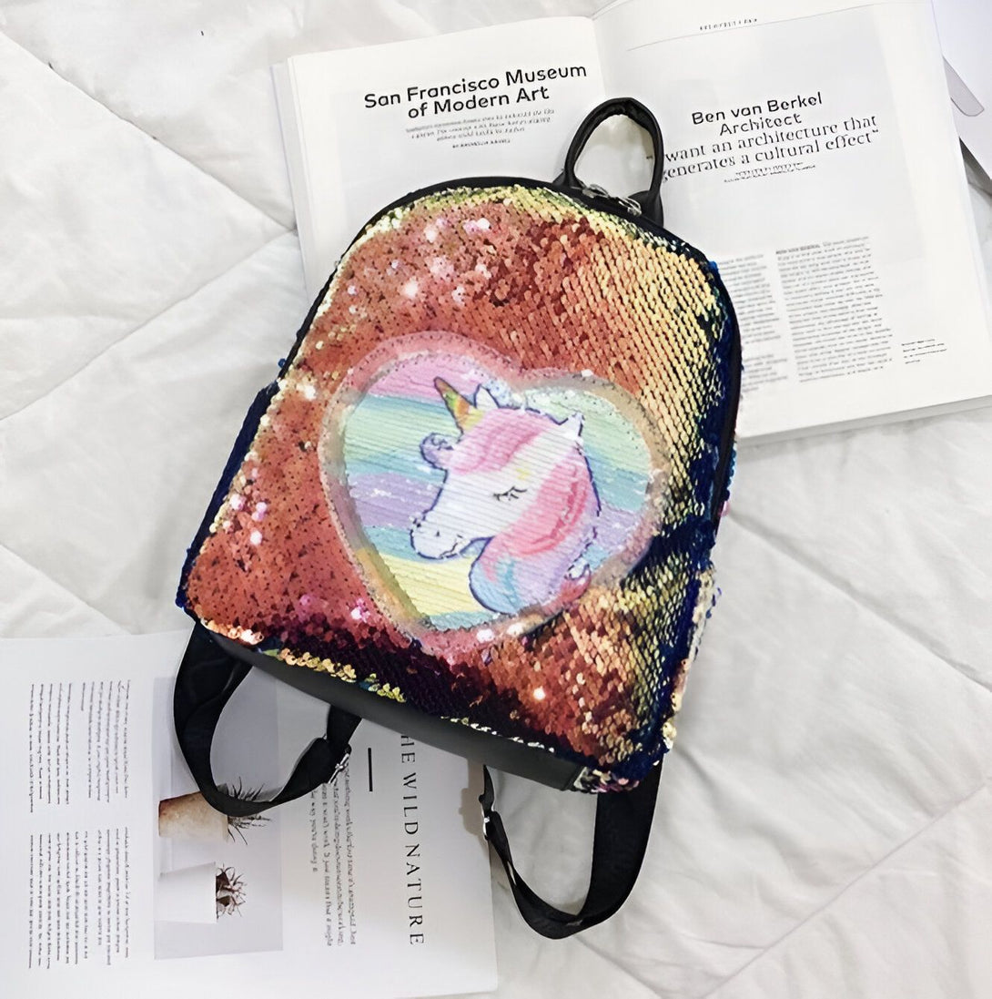 Une image d'un sac à dos à paillettes avec une image de licorne. Le sac à dos est de couleur arc-en-ciel et la licorne est de couleur rose et blanche.