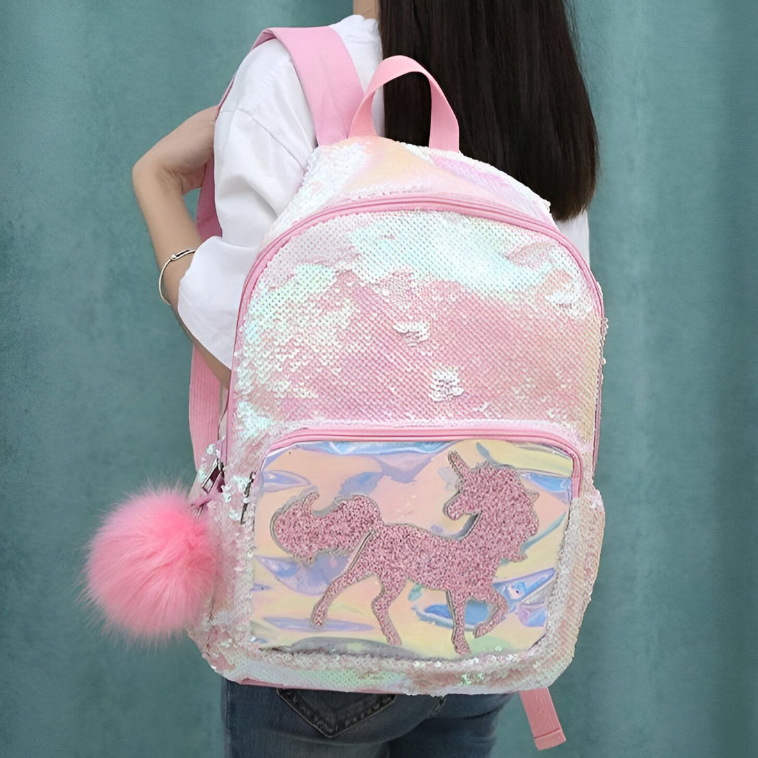 Une jeune fille porte un sac à dos rose à paillettes avec une image de licorne. Le sac à dos est porté sur les épaules de la fille et a une fermeture à glissière. La fille porte également un bracelet rose au poignet droit.