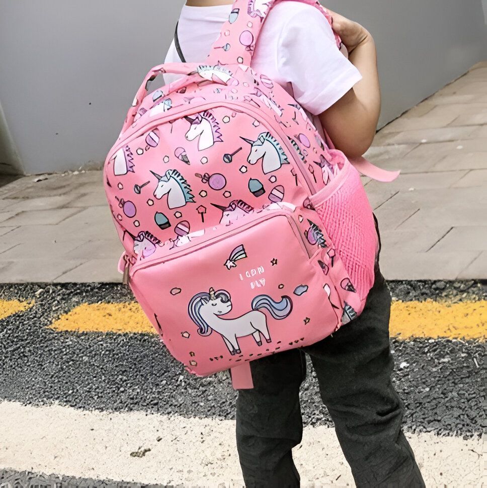 Une petite fille porte un sac à dos rose avec une image de licorne dessus. Le sac à dos a une poche latérale en filet et des bretelles réglables. La petite fille porte également un pantalon noir et une chemise rose.