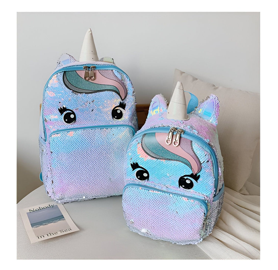 Deux sacs à dos à paillettes en forme de licorne sont placés sur un lit. Le grand sac à dos est bleu clair avec une crinière et une queue arc-en-ciel, et le petit sac à dos est rose avec une crinière et une queue violettes. Les deux sacs à dos ont des yeux et une bouche brodés.