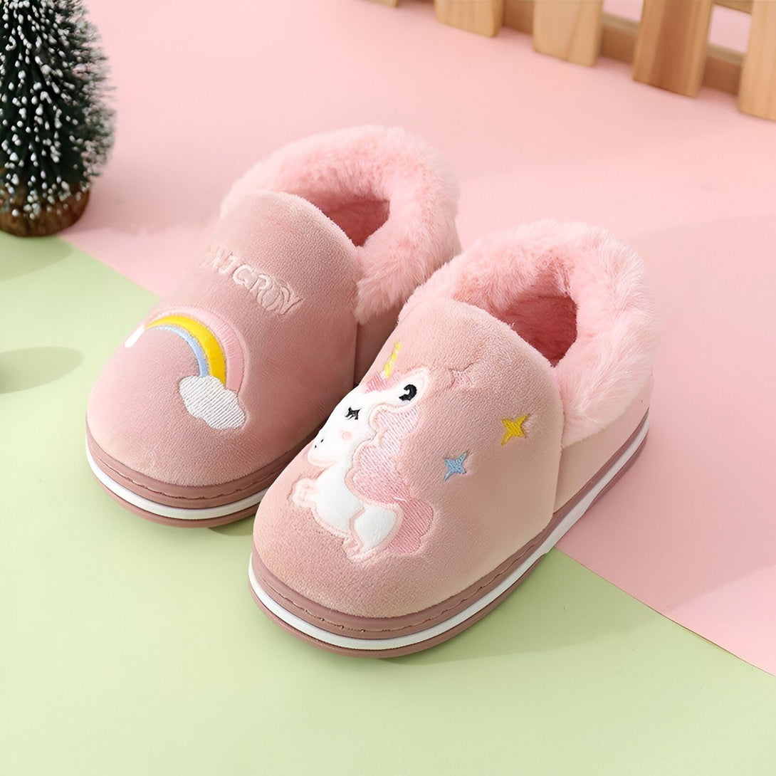 Une paire de pantoufles roses pour enfant avec une licorne et un arc-en-ciel brodés sur le dessus. Les pantoufles sont bordées de fourrure blanche.