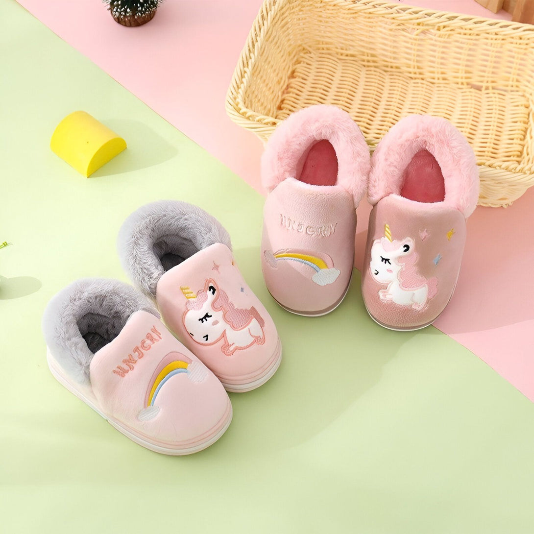 Une paire de chaussons licorne rose et grise pour enfant. Les chaussons sont brodés d'une licorne et d'un arc-en-ciel. Ils sont doublés de fourrure pour tenir les pieds au chaud.