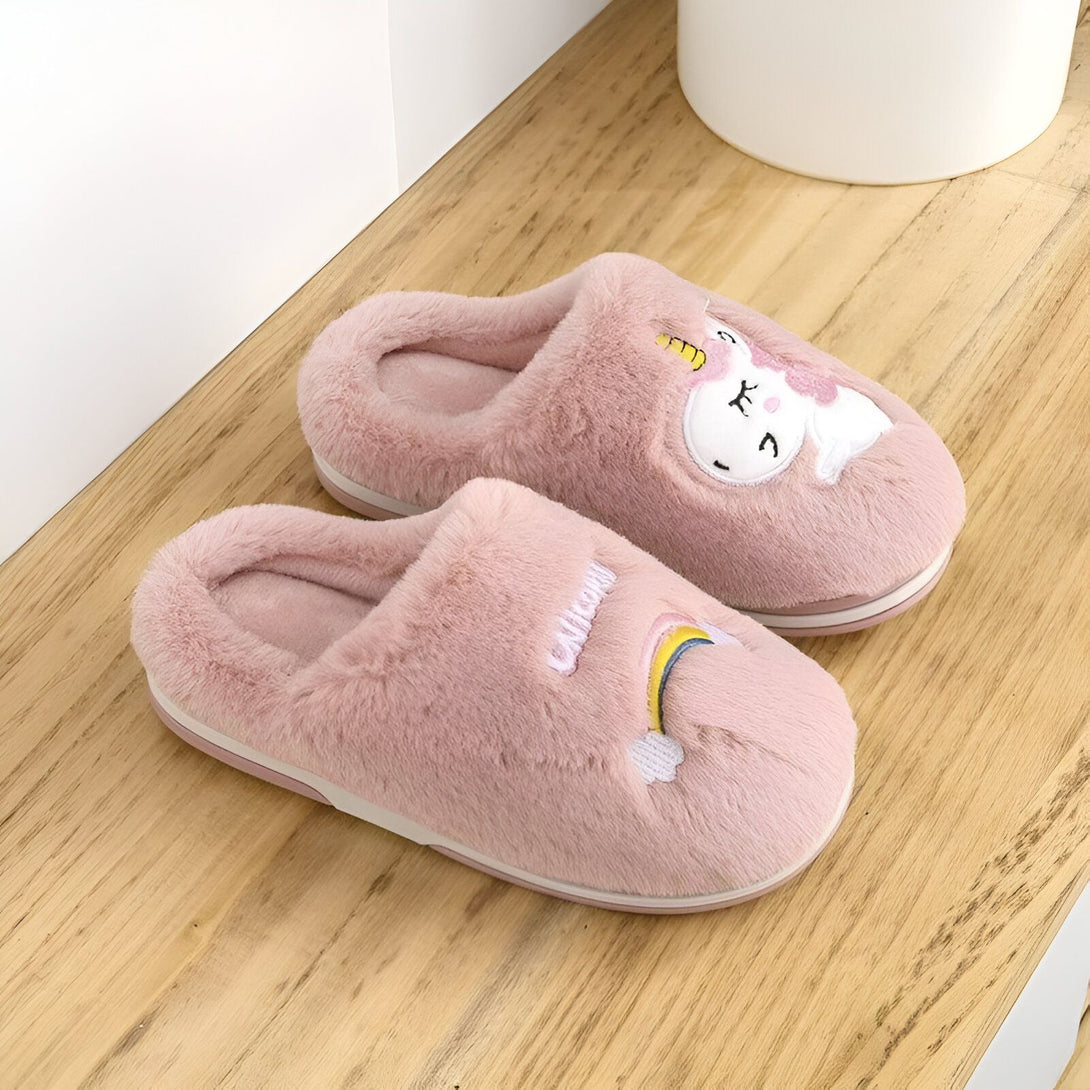 Une paire de pantoufles licorne en peluche rose avec une corne et un arc-en-ciel brodés sur le dessus. Les pantoufles sont doublées de fourrure blanche et ont une semelle antidérapante.