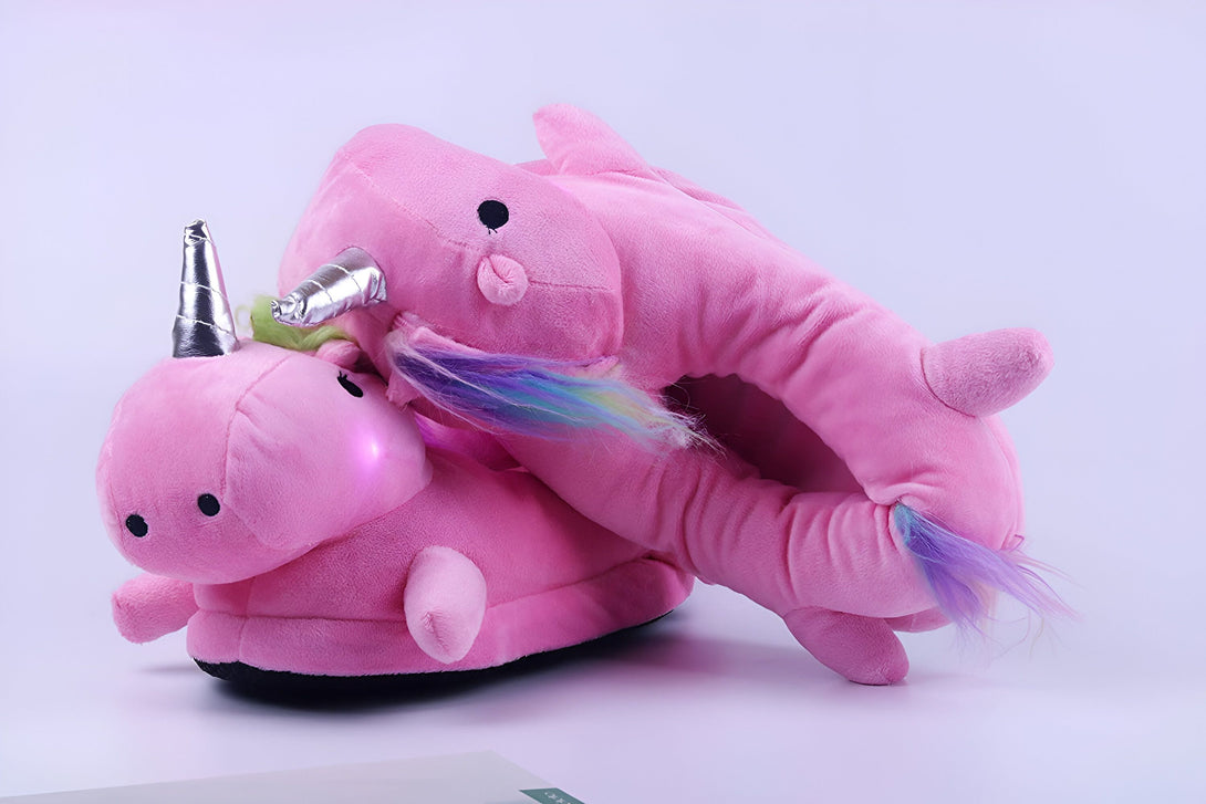 Une paire de pantoufles licorne en peluche rose avec une corne et une queue arc-en-ciel. Les pantoufles sont éclairées par des LED.