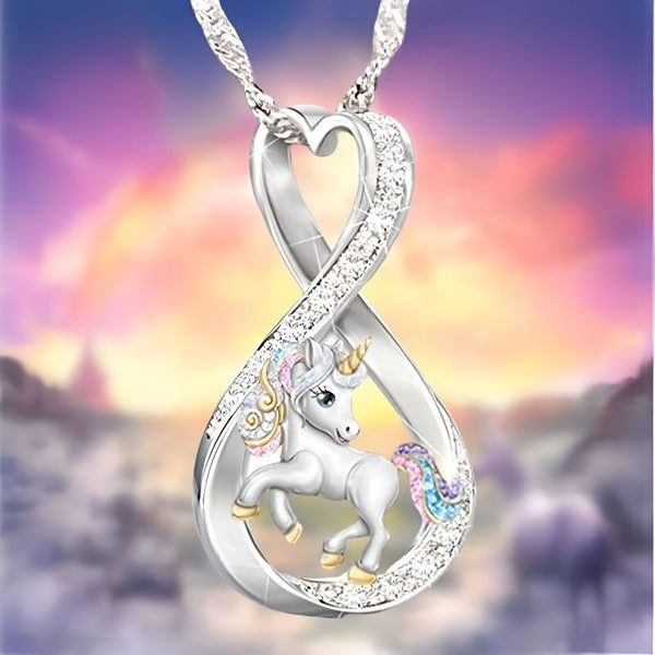 L'image représente un pendentif en forme de cœur avec une licorne. La licorne est blanche avec une crinière et une queue arc-en-ciel. Le pendentif est en argent et serti de strass.