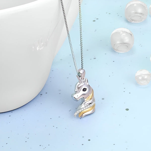 L'image représente un pendentif en forme de tête de licorne. La licorne est représentée de profil et porte une crinière et une corne dorées. Le pendentif est en argent et est suspendu à une chaîne en argent.