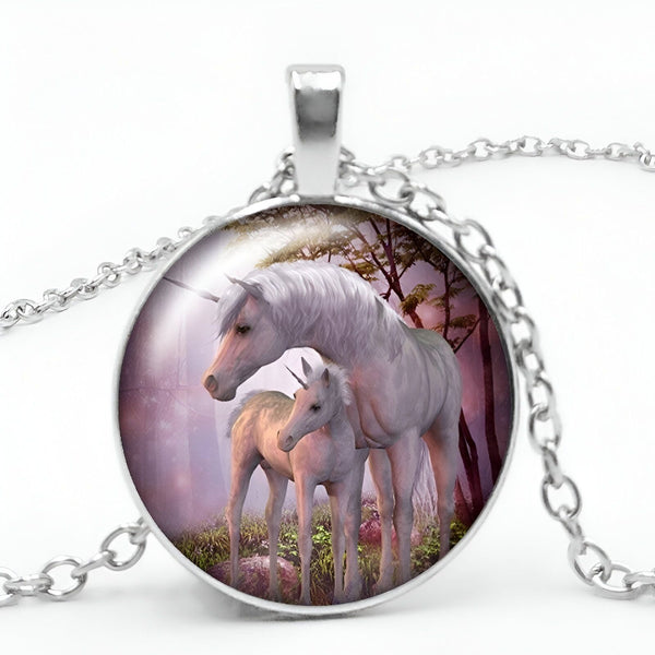 Un pendentif rond en métal argenté avec une chaîne. Le pendentif représente une licorne blanche et son petit dans une forêt. La licorne et son petit sont debout sur un sol vert avec des arbres et des montagnes en arrière-plan. Le ciel est rose et il y a un arc-en-ciel qui traverse le ciel.