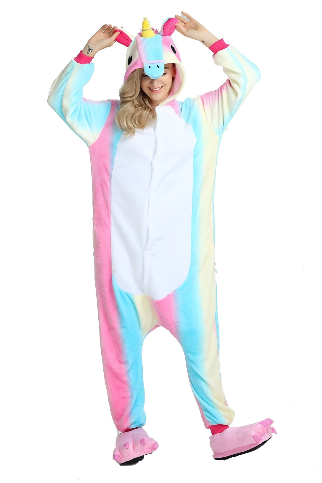 Une femme porte une combinaison pyjama licorne arc-en-ciel. La combinaison est rose, bleue et blanche. La femme a les cheveux blonds et elle sourit. Elle porte des chaussettes licorne roses.