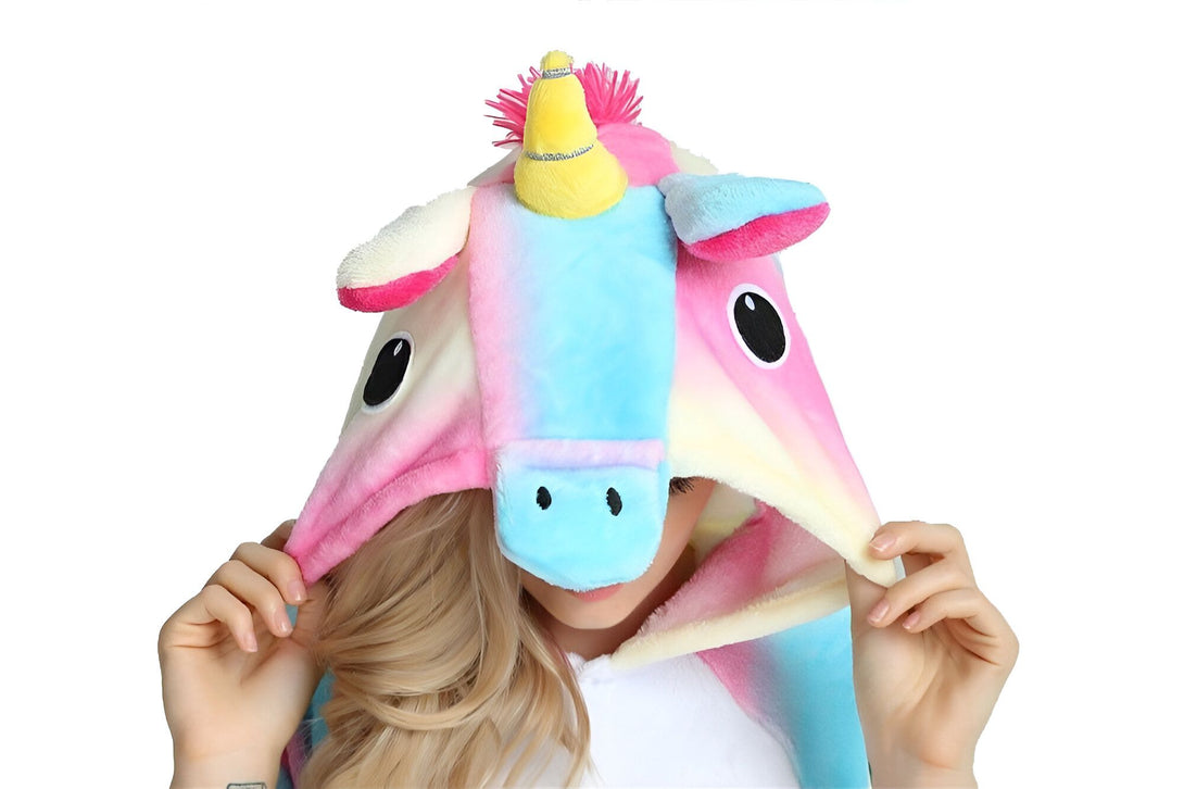 Une femme porte une combinaison pyjama licorne. La combinaison est rose, bleue et blanche avec une capuche licorne. La femme a les yeux bleus et les cheveux blonds. Elle sourit et regarde l'appareil photo.