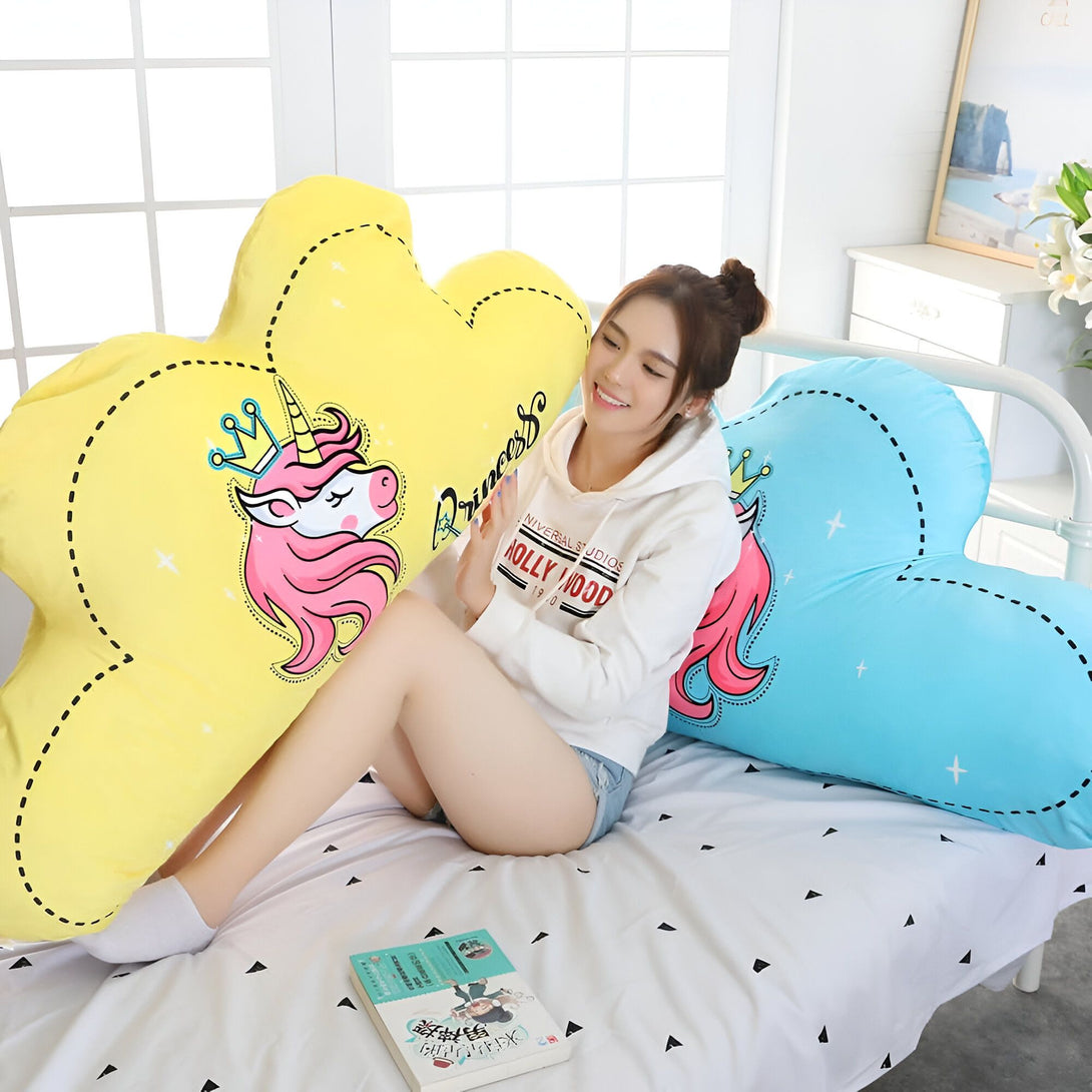 Femme souriante assise sur un lit avec coussins en forme de nuage décorés de licornes. Oreillers moelleux jaunes et bleus avec motif princesse. Ambiance douillette et rêveuse., 45501049569535 | Licorne Shop