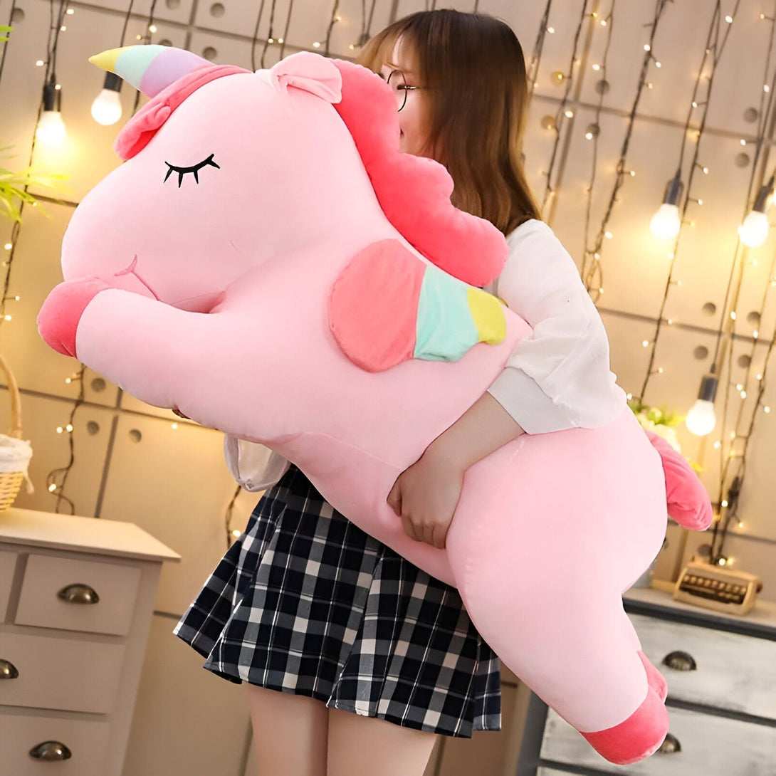 Une femme tenant une grande peluche licorne rose avec une crinière arc-en-ciel. La licorne a des yeux brodés et un corps doux. Des lumières féériques sont en arrière-plan., 45501065986303 | Licorne Shop