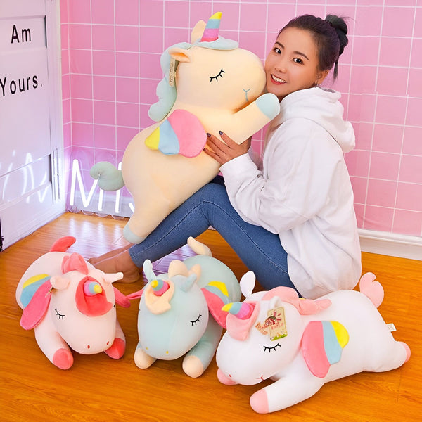 Une femme souriante serre un grand doudou licorne jaune. Autour d'elle, plusieurs autres peluches licornes colorées sont posées sur le sol. Décor rose et inscription lumineuse en arrière-plan., 45501048684799 | Licorne Shop