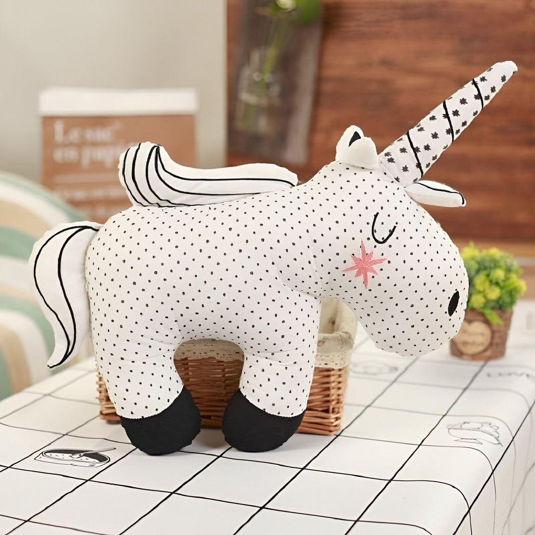 Peluche licorne blanche à pois noirs avec une crinière et une queue rayées. Une étoile rose orne sa joue et son unique corne est décorée de motifs noirs. Posée sur une table à carreaux dans un intérieur lumineux., 45501060382975 | Licorne Shop