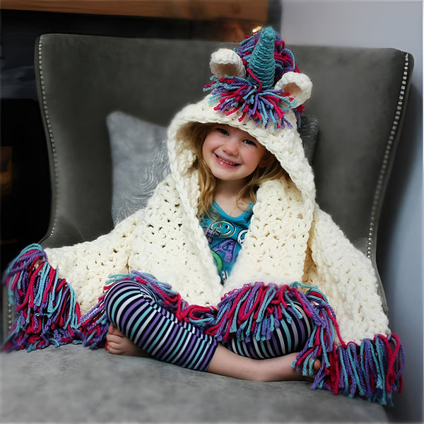 Une fillette souriante assise dans un fauteuil, enveloppée dans une couverture à capuche blanche en forme de licorne. La couverture est ornée de franges colorées en bleu, rose et violet. Elle porte un legging rayé et un t-shirt bleu., 45502087364863 | Licorne Shop