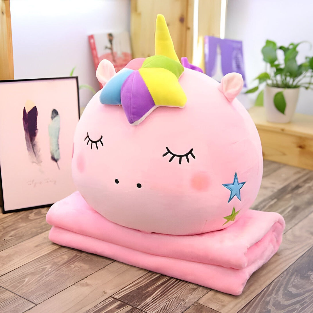 Image d'un coussin en peluche en forme de tête de licorne rose avec une corne multicolore, les yeux fermés et une étoile brodée, posé sur une couverture polaire rose pliée. Décoration intérieure visible en arrière-plan., 45501048520959 | Licorne Shop