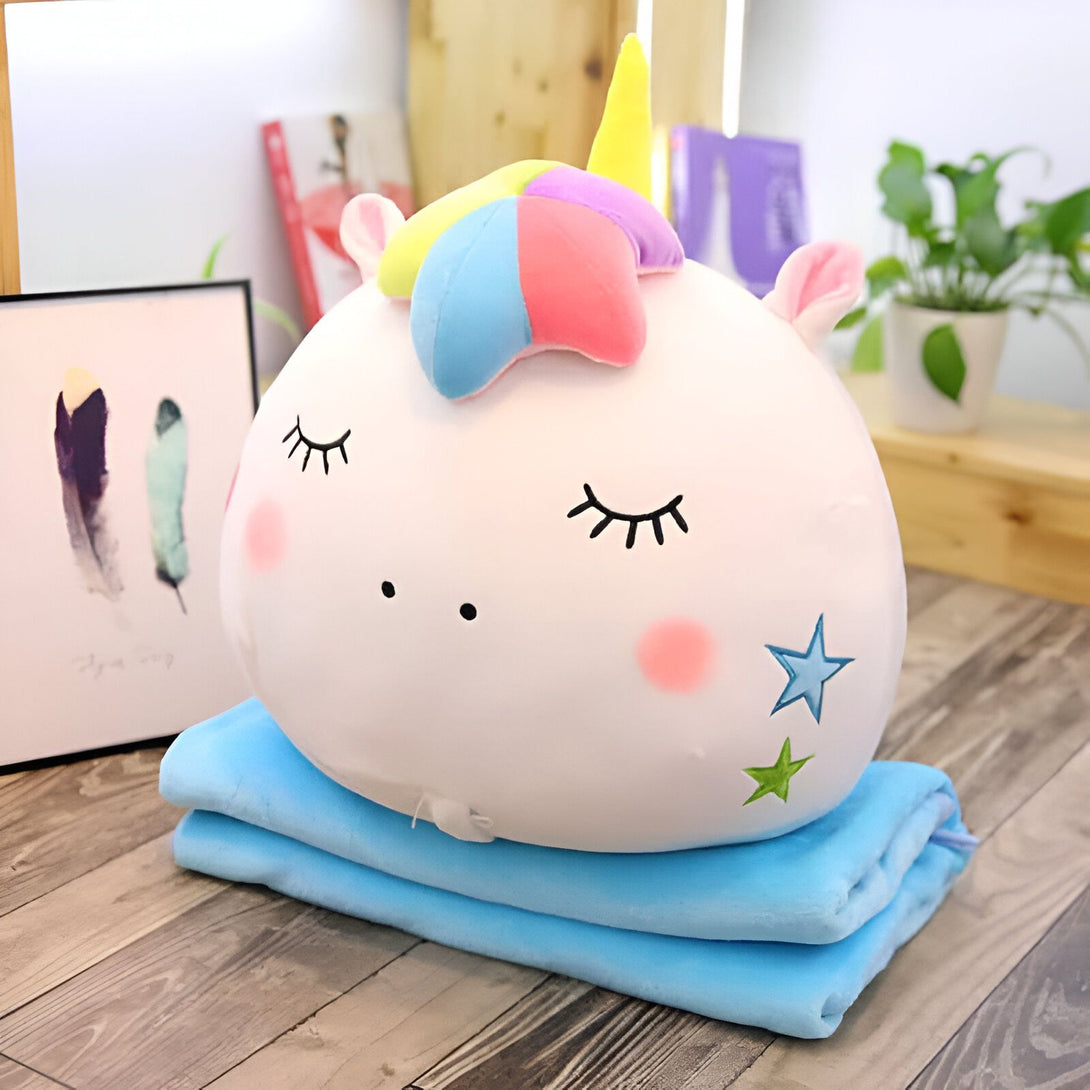 Peluche licorne ronde avec crinière arc-en-ciel et joues roses reposant sur une couverture bleue, avec des décorations floues en arrière-plan., 45501048783103 | Licorne Shop