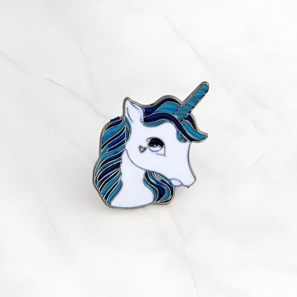 Pin's en émail représentant la tête d'une licorne blanche avec une crinière et une corne bleues, avec un petit cœur dessiné à la place de l'œil., 45502390894847 | Licorne Shop