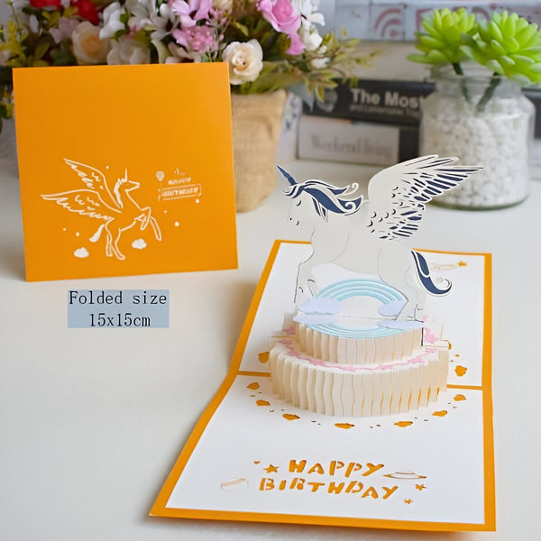 Carte d'anniversaire pop-up. Design élaboré avec un gâteau à étages et un décor d'unicorne ailée. L'image capture une carte ouverte montrant les détails découpés et un fond de fleurs et de livres, créant une ambiance festive et décorative., 45501112910079 | Licorne Shop